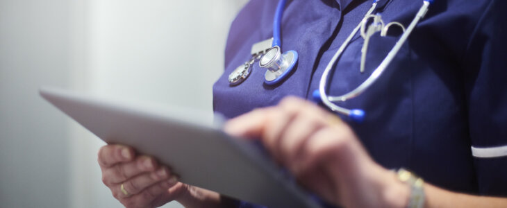Female surgeon typing on digital tablet in hospital or surgery. She is wearing a dark blue nurse’s top and has her stethoscope around her neck. She is looking at her patients records on her digital tablet and sending emails.